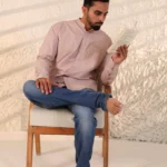 Man in blush pink shirt and blue jeans sitting on a wooden stool.