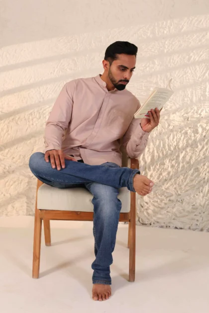Man in blush pink shirt and blue jeans sitting on a wooden stool.