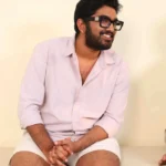 Man in light pink shirt smiling, seated with hands behind him.