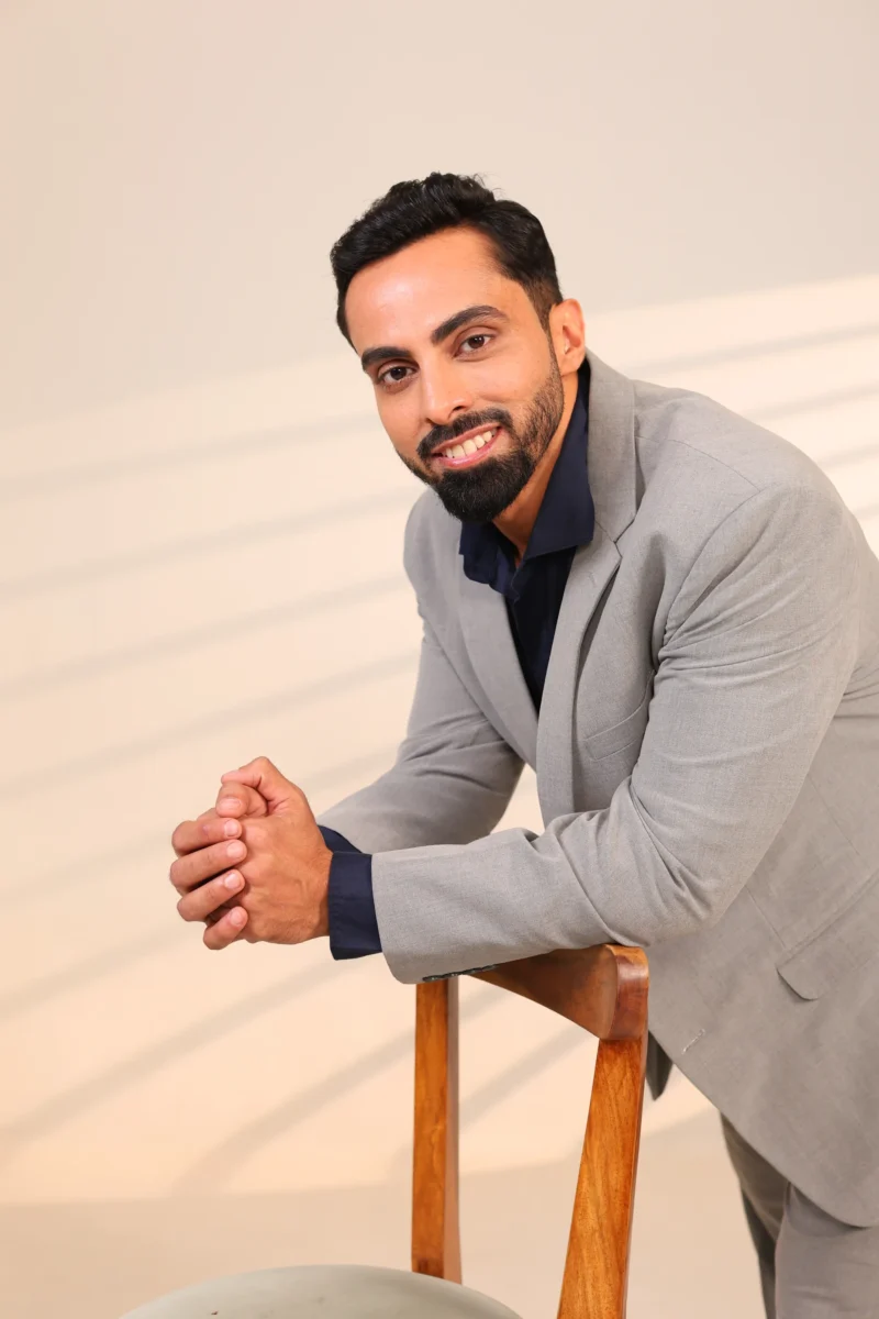 Man in grey blazer and navy shirt facing right with hands together.